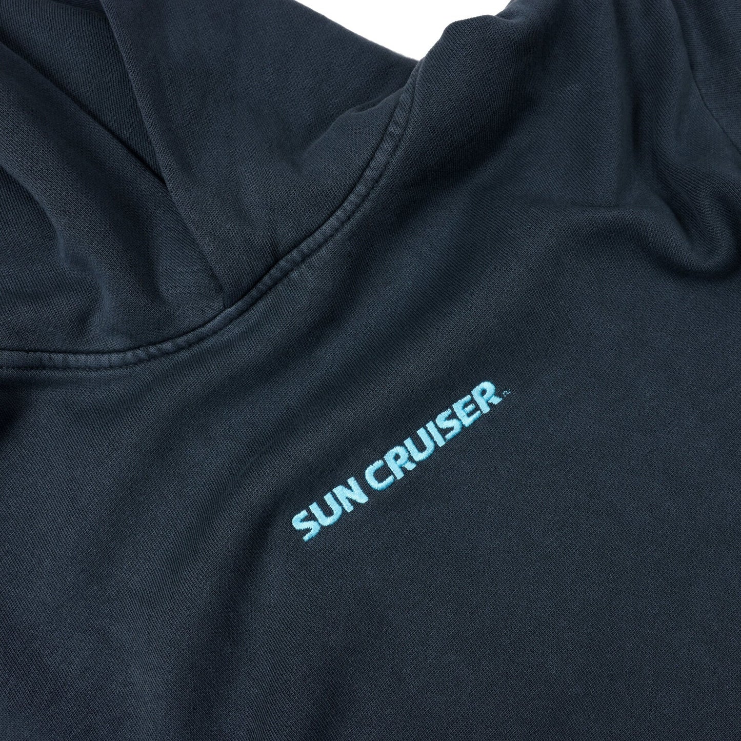A close-up of the chest area of a dark blue, faded hooded sweatshirt. The words 'SUN CRUISER' are printed in light blue, slightly faded text, running diagonally across the fabric from the left shoulder towards the center. The stitching around the hood is visible.