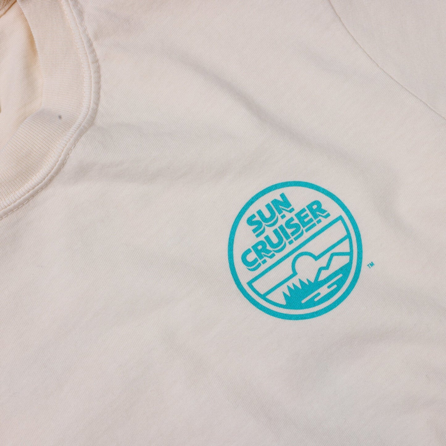 A close-up of the left chest area of a cream-colored t-shirt, showing a circular logo in teal. The logo features the words 'SUN CRUISER' in a stacked format at the top, and a stylized landscape below with a sun, mountains, and water, all within the teal circle.