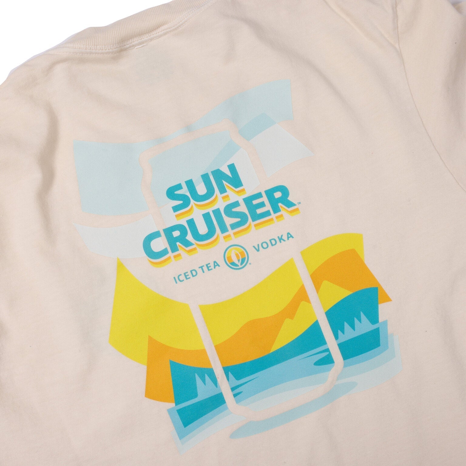 A close-up of the upper back of a cream-colored t-shirt, revealing a large, colorful graphic. The graphic is a stylized depiction of a sun, mountains, and water scene with layers of yellow, orange, and blue. The words 'SUN CRUISER' are prominently displayed in blue text within the graphic, with 'ICED TEA' on the left and 'VODKA' on the right in smaller text below it, separated by a small leaf-like logo.