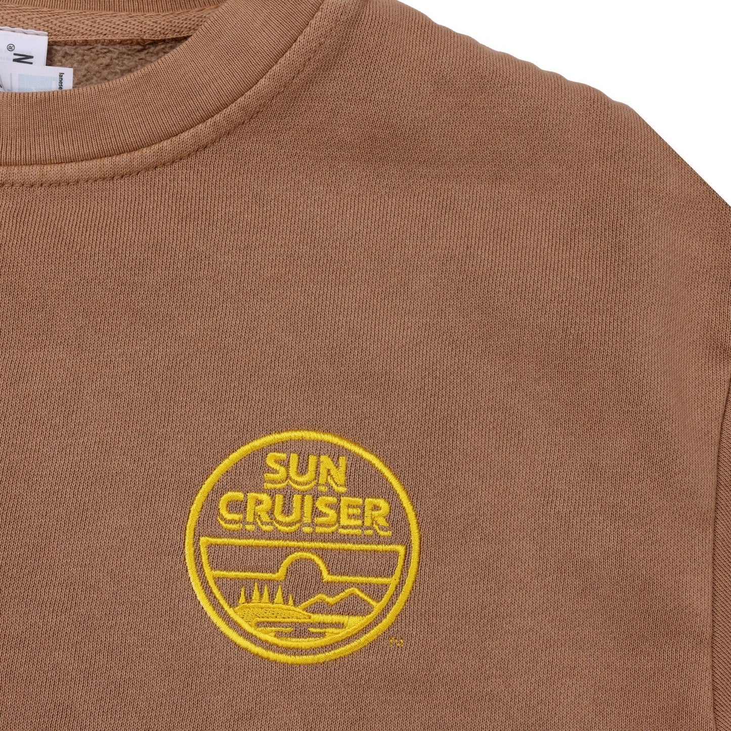 A close-up of the chest area of a light brown crewneck sweatshirt, showing a circular embroidered logo in yellow. The logo features the words 'SUN CRUISER' stacked at the top, and a graphic below depicting a landscape with mountains, water, and trees, all outlined in yellow. 