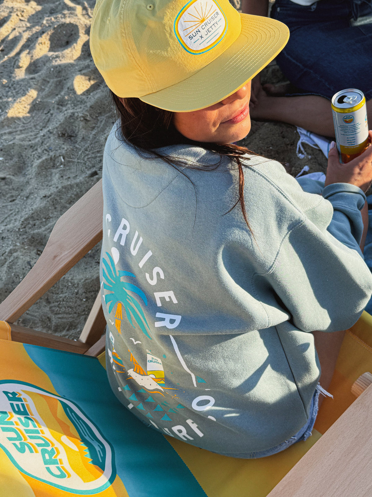 Person sitting on a beach chair wearing a yellow cap and gray hoodie with 'Sun Cruisers' branding.