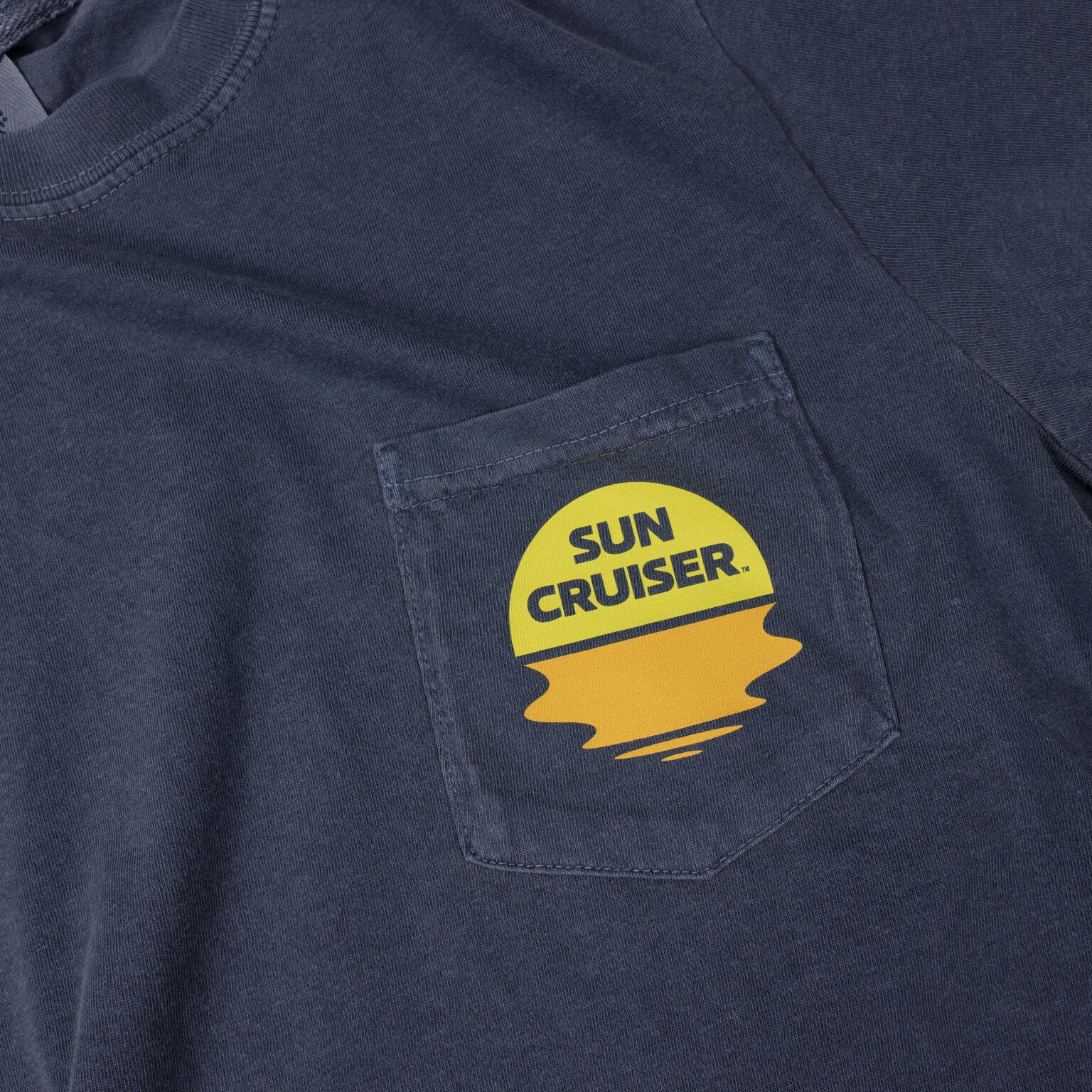 A close-up of the left chest pocket on a faded blue t-shirt. On the pocket, there is a round yellow and orange graphic resembling a sunset over water. The words 'SUN CRUISER' are printed in blue on the yellow part of the graphic. The stitching of the pocket is visible.