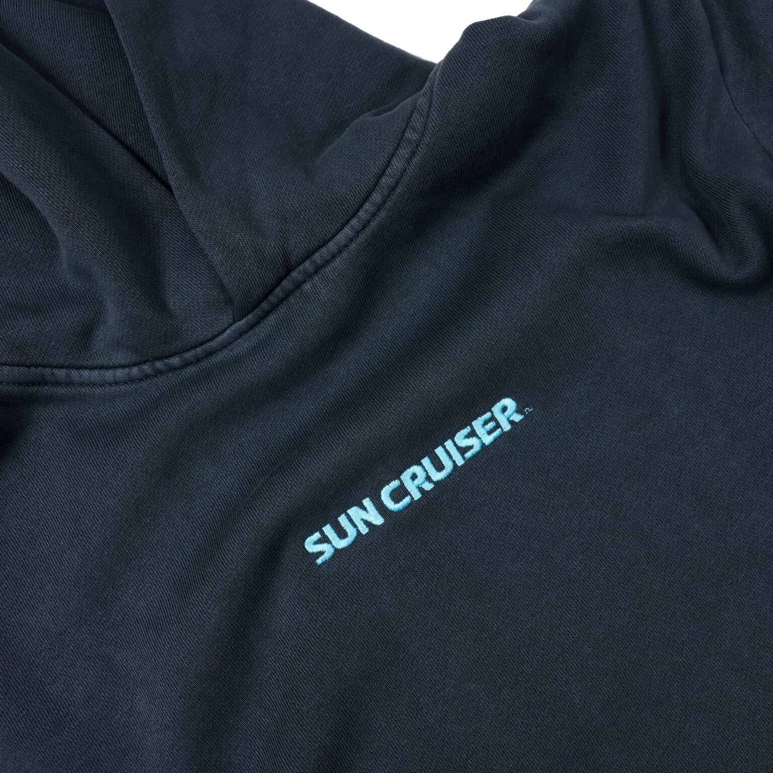 A close-up of the chest area of a dark blue, faded hooded sweatshirt. The words 'SUN CRUISER' are printed in light blue, slightly faded text, running diagonally across the fabric from the left shoulder towards the center. The stitching around the hood is visible.