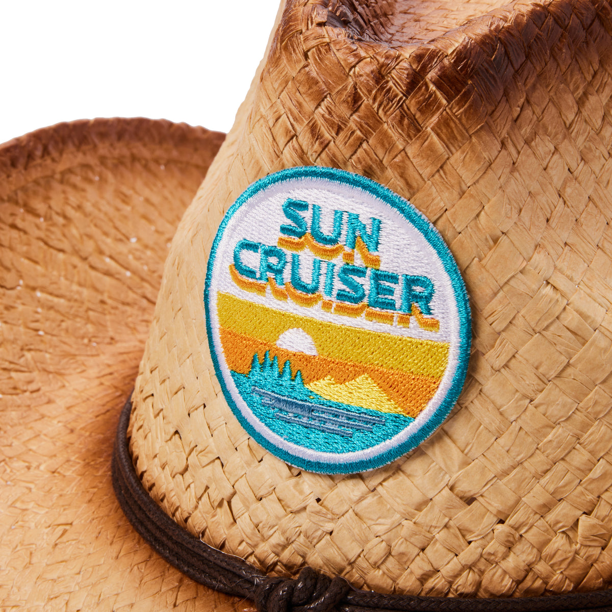 Close-up of a woven straw cowboy hat with a round embroidered patch reading “SUN CRUISER” on the brim. The patch features a turquoise, yellow, and white sunset design.
