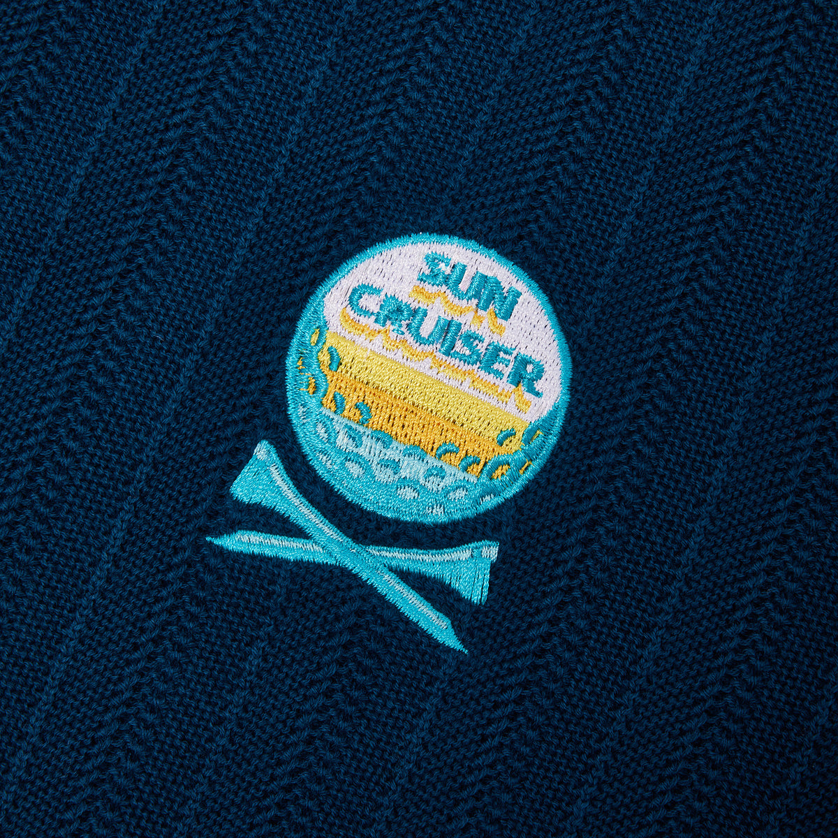 Close-up of a navy blue textured fabric with a circular embroidered patch reading 'SUN CRUISER' in blue and yellow, plus crossed teal lines below.