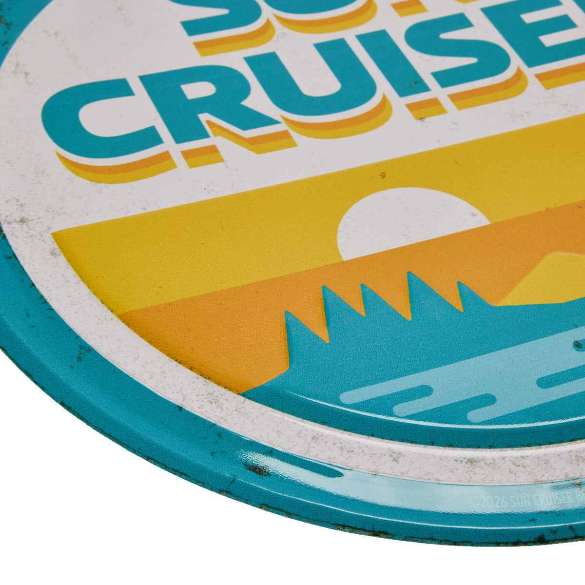 Close-up of a vintage-style metal sign with the words "SUN CRUISER" in large teal letters over a yellow and orange sunset graphic with a white sun and blue mountains.