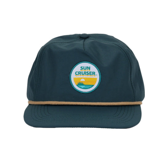 A navy blue hat with a flat brim and a tan rope accent above the brim. On the front, there is a circular patch with a blue border and a white inner circle. Inside the white circle, 'SUN CRUISER' is written in yellow, above a yellow and blue graphic depicting a sunset over water with a small.