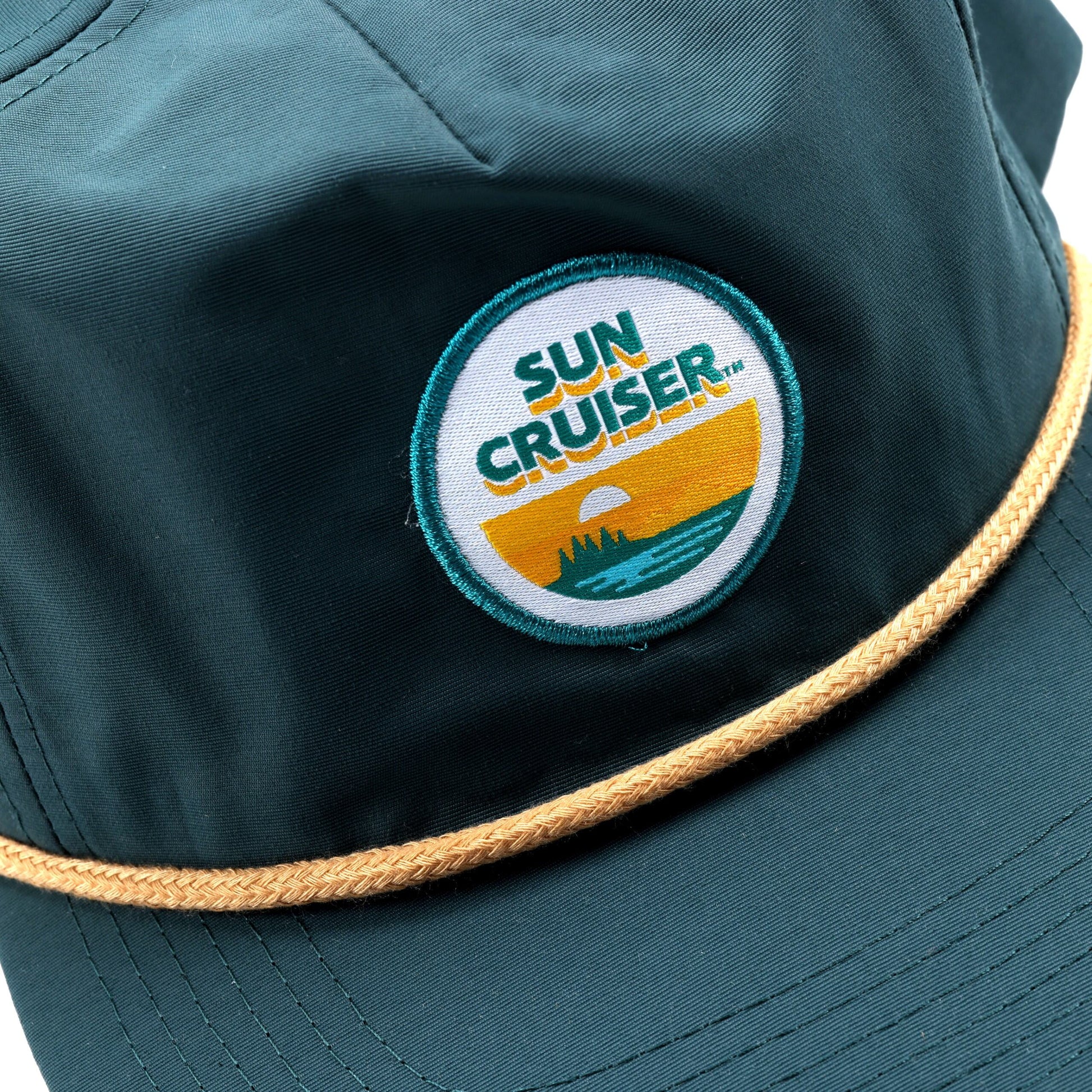A close-up view of a navy blue hat. On the front, there is a circular embroidered patch with a blue border. Inside the patch, the words 'SUN CRUISER' are in a blue font over a yellow band, which is above a teal graphic depicting a sunset over water. A tan rope detail runs across the brim of the cap.