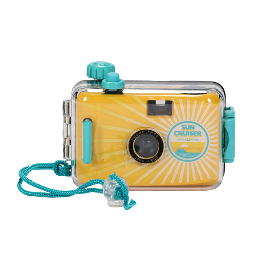 Front view of a yellow disposable camera with a sunburst pattern, housed in a clear waterproof case. The camera's black lens is centered and labeled 'FOCUS FREE 28mm LENS'. On the right, a circular 'SUN CRUISER' logo depicts a sun, waves, and mountains. The clear case has turquoise accents, including a winding knob, shutter button, and side latches. A turquoise braided wrist strap with a clear stopper is attached.