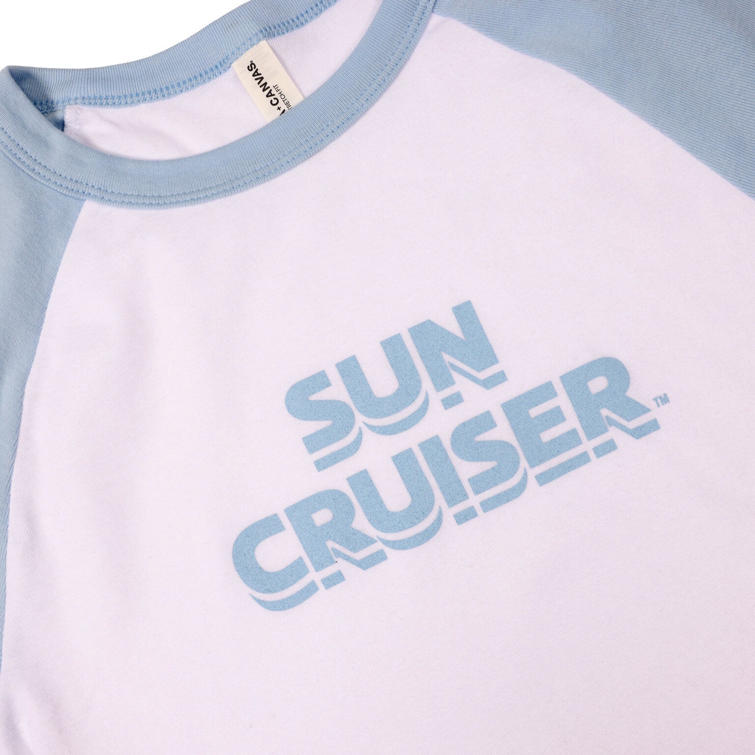 A close-up of a white cropped t-shirt with light blue raglan sleeves and collar. The chest area prominently displays the words 'SUN CRUISER' in a bold, light blue, block font with a slight shadow effect. A portion of the 'BELLA+CANVAS' brand tag is visible near the collar.