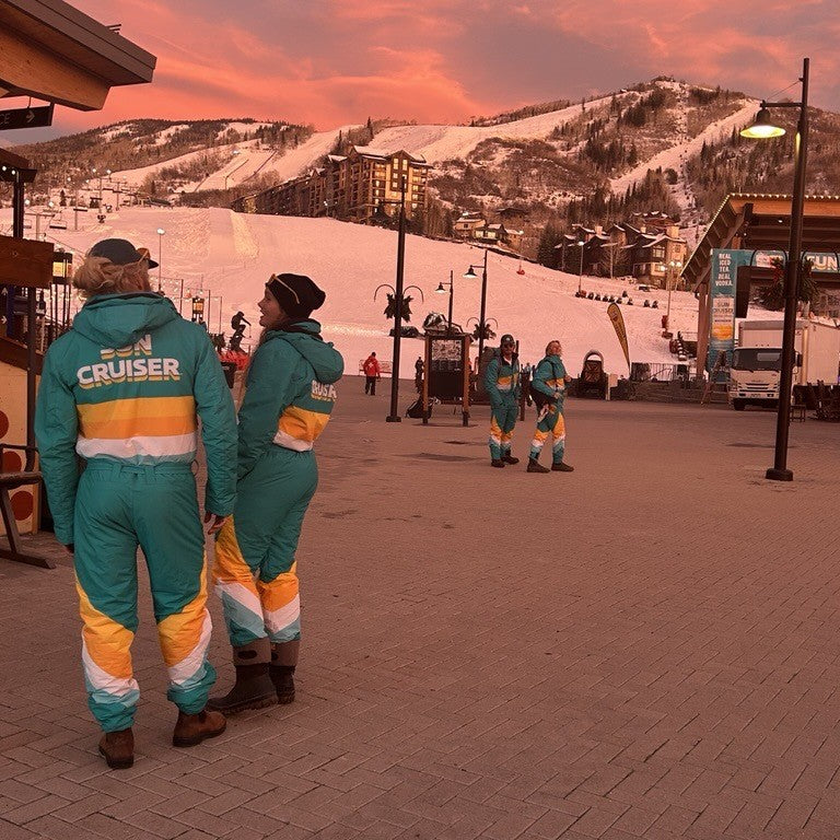 Two people in matching teal and orange ski uniforms standing on a snowy mountain slope with a sunset sky.