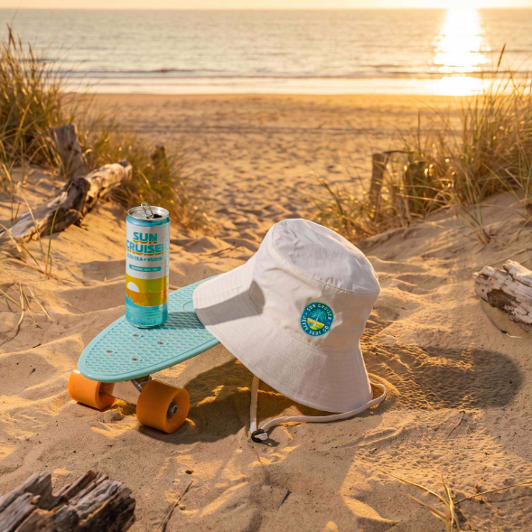 A white wide-brim sun hat with a blue and yellow circular logo sits on a sandy beach next to a light blue penny board with orange wheels. A can of Sun Cruiser is placed on top of the board, with the ocean and golden sunset in the background.