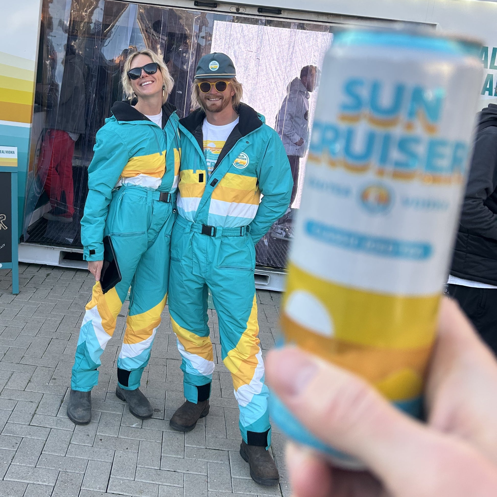 Two people in matching teal and yellow outfits standing next to a can of Sun Cruiser.