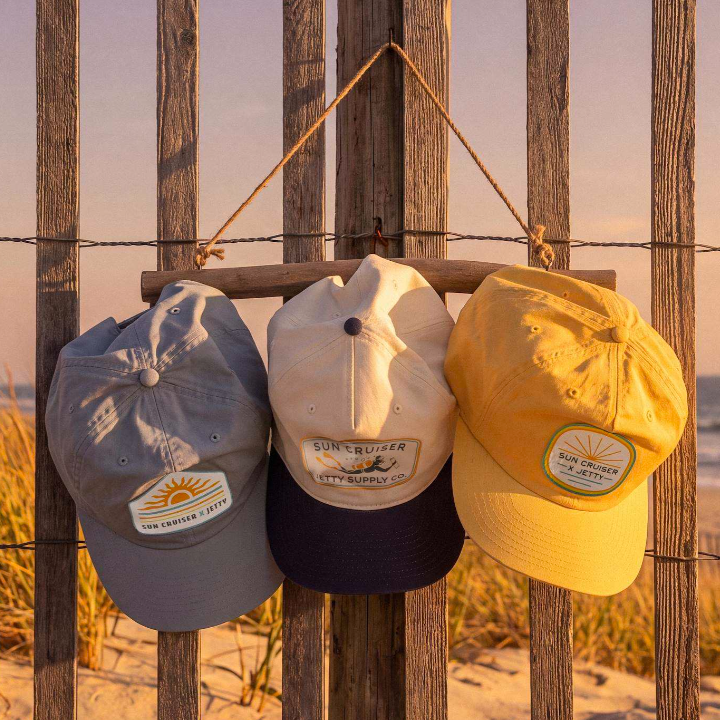 Three baseball caps hanging from a rope on a wooden beach fence at sunset: a gray cap on the left, a cream cap with a dark navy brim in the center, and a mustard yellow cap on the right. Each cap has a patch logo reading 'Sun Cruiser x Jetty'.