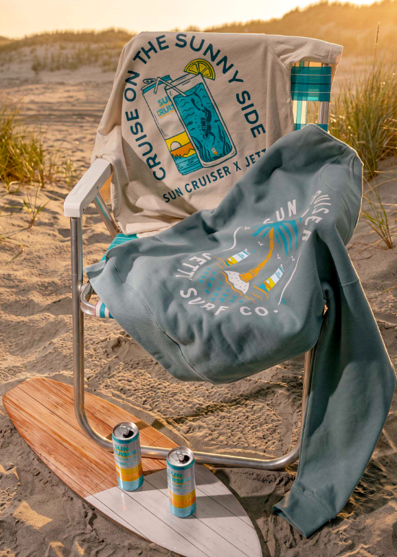 A beach chair set on sand at sunset with a large beige sweatshirt draped over the back and a slate blue sweatshirt spread across the seat. The shirts feature a tropical cocktail graphic and the text 'Cruise on the Sunny Side' and 'Sun Cruiser x Jetty Surf Co', with two cans resting on a surfboard nearby.